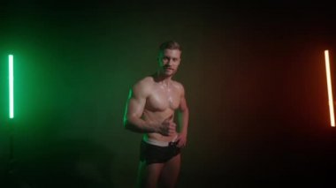Çıplak vücutlu bir sporcu kameraya bakıyor ve parmağını yukarı kaldırıyor. Gülümsüyor. Kamera yakınlaştırıyor. Turuncu ve yeşil ışık onun üzerinde parlıyor. 4K