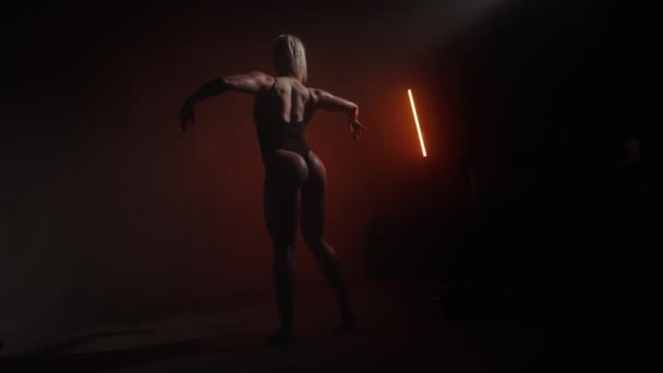 一个穿着紧身衣的漂亮的健身模特正在摆姿势.她在转身离去。她在炫耀后背和臀部的肌肉。她身上闪烁着橙色的光芒.4K — 图库视频影像