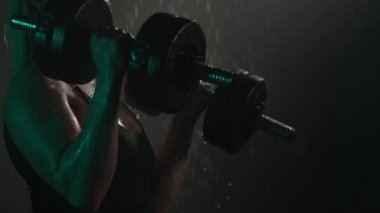 Fitness modeli duşta duruyor ve pazı egzersizi yapıyor. O, kollarını halterlerle büküyor. Kamera dambıllara odaklanıyor. Vücudundan su akıyor. 4K