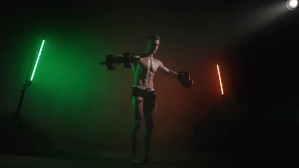 En idrottsman svänger hantlar åt sidorna. Han gör en axelövning. Se hur hans muskler fungerar. Ett orange och grönt ljus lyser på honom. 4K — Stockvideo