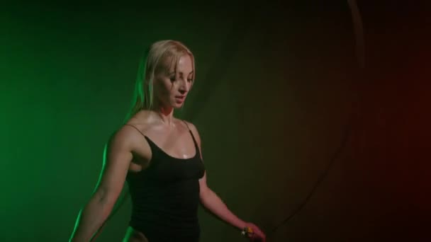 ボディスーツのフィットネスモデルがロープを飛び回っています。彼女はロープを交差させている。心臓訓練だ。オレンジと緑の光が彼女の上に輝いている。4K 50fps — ストック動画