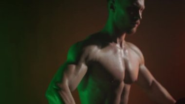 Sporcu halterleri yana doğru sallar. O bir omuz egzersizi yapıyor. Kaslarının nasıl çalıştığına bak. Kamera dambıllara odaklanıyor. Turuncu ve yeşil ışık onun üzerinde parlıyor. 4K