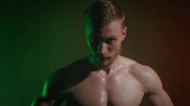 Çıplak vücutlu genç bir sporcunun portresi. Başını kaldırıyor ve kameraya bakıyor. Turuncu ve yeşil ışık onun üzerinde parlıyor. 4K