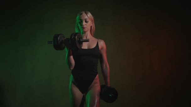 O modelo de fitness está de pé e fazendo um exercício de bíceps. Ela está dobrando o braço com um halteres. Ela está a olhar para a câmara. Uma luz laranja e verde está brilhando sobre ela. 4K 50fps — Vídeo de Stock