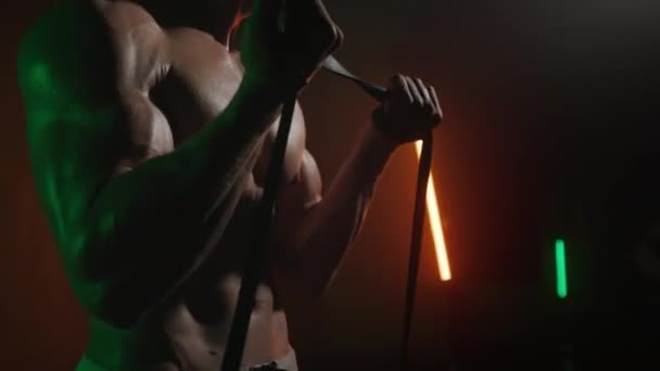 The sportsman is standing and performing a bicep exercise. He is lifting the resistance bands. The camera is focusing on his body. An orange and green light is shining on his. 4K 50fps — Stock Video