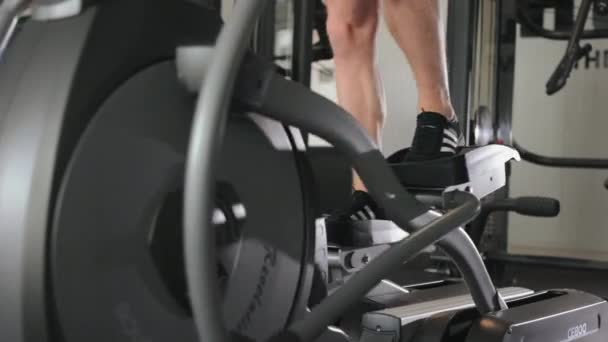 En sporty mann er engasjert på en elliptisk trener. Kameraet fokuserer på beina hans. Cardio trening. Trene i gymsalen. 4K – stockvideo