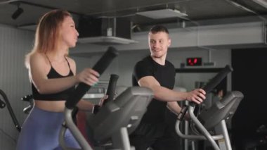 Bir adam ve bir kız eliptik bir eğitmen üzerinde çalışıyorlar. Konuşuyorlar ve gülümsüyorlar. Kardiyo eğitimi. 4K