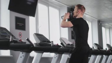 Sporcu, su içiyor. Spor salonundaki koşu bandında yürüyor. Spor salonunda antrenman. O, pencereden dışarı bakıyor. 4K