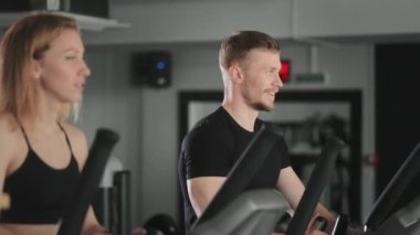 Bir adam ve bir kız eliptik bir eğitmen üzerinde çalışıyorlar. Bir adam gülümsüyor. Kardiyo eğitimi. Kamera onların ellerine odaklanmış durumda. 4K