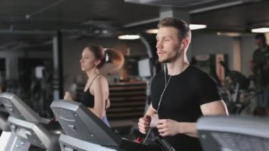 Genç bir atletik adam koşu bandında koşuyor ve kulaklıkla müzik dinliyor. Yanında bir kadın koşuyor. Spor salonundalar. 4K