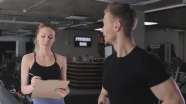 Bir adam koşu bandında koşuyor. Bir spor koçu onun yanında duruyor ve performansını kontrol ediyor. Bir belgeye veri yazıyor. 4K