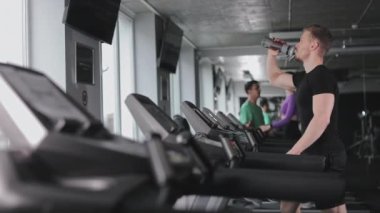 Sporcu, su içiyor. Spor salonundaki koşu bandında yürüyor. Uzun kardiyo seansları. Spor salonunda antrenman. 4K