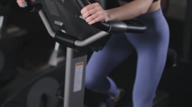 Genç bir atletik kadın egzersiz bisikletine biniyor. O tayt giyiyor. Spor salonunda kardiyo eğitimi. 4K