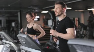 Genç bir atletik adam koşu bandında koşuyor ve kulaklıkla müzik dinliyor. Yanında bir kadın koşuyor. Gülümsüyorlar ve konuşuyorlar. 4K