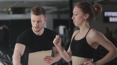 Genç bir kadın koşu bandında koşuyor. Bir spor koçu onun yanında duruyor ve performansını kontrol ediyor. Bir koşu bandı çalışma programı seçiyor. 4K