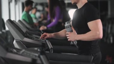 Bir adam koşu bandında yürüyor ve bir eğitim programı seçiyor. O, elinde bir şişe su tutuyor. Spor salonunda antrenman. 4K