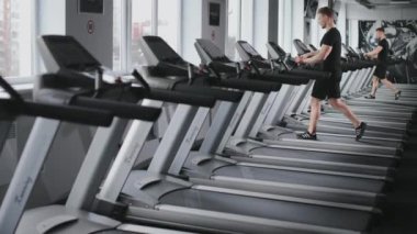 Sportif bir adam koşu bandında yürüyor. Bir eğitim programı seçiyor. Spor salonunda kardiyo eğitimi. 4K