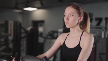 Egzersiz bisikletine binmiş genç bir atletik kadının portresi. Spor salonunda kardiyo eğitimi. Kamera yukarıdan aşağıya hareket ediyor. 4K