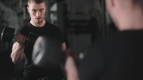 Egy sportos ember bicepszgyakorlatot végez. Ott áll a tükör előtt, és figyeli a gyakorlat helyes végrehajtását. A kamera alulról felfelé mozog. 4K — Stock videók