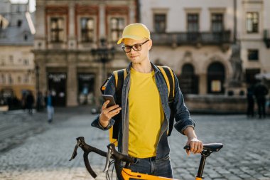 Mutlu bir teslimatçı bisiklette oturuyor ve akıllı telefonuyla mesaj atıyor. Omzunda sarı bir sırt çantası taşıyor..