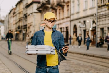 Genç, mutlu bir teslimatçı ayakta ve elinde üç pizza kutusu tutuyor. O, akıllı telefonda gülümsüyor ve mesaj atıyor. Sarı bir şapka ve gözlük takıyor. Arka planda eski şehrin merkezi.