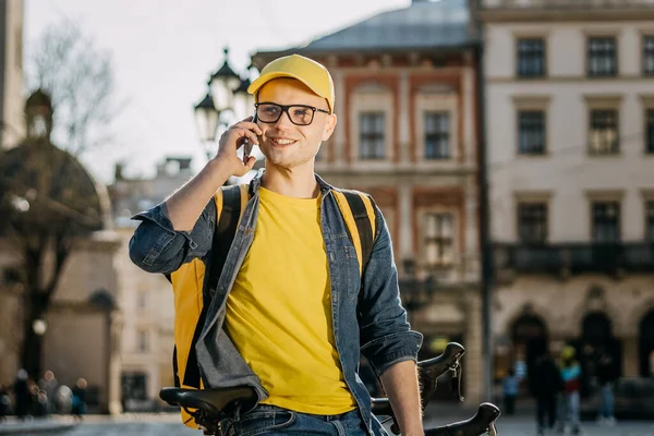 Bisiklette oturan ve telefonda duygusal olarak konuşan genç bir teslimatçının portresi. Omuzlarında sarı bir sırt çantası taşıyor..