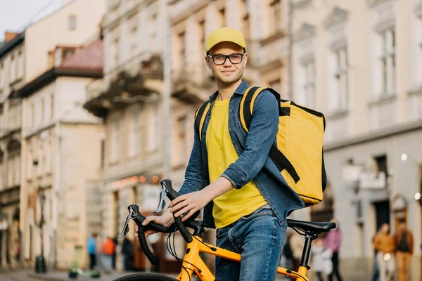 Portrait d'un livreur heureux assis sur un vélo et regardant la caméra. Il porte un sac à dos jaune sur ses épaules et sa casquette. — Photo