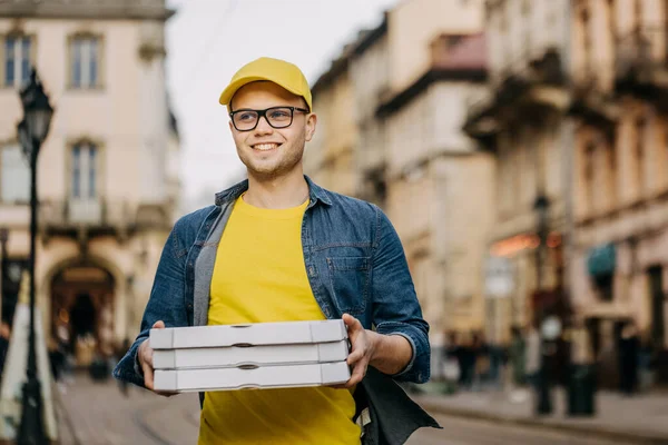 Genç, mutlu bir teslimatçı üç pizza getiriyor. Gülümsüyor. Sarı bir şapka ve gözlük takıyor. Eski şehrin merkezine doğru gidiyor..