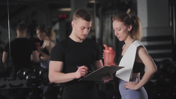 The trainer is discussing post-workout performance with his client. He is holding a folder with tests. A woman is holding a bottle of water and a towel. They are standing in the gym. 4K — 图库视频影像