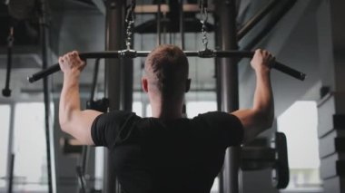 Sporcu bir blok makinesinin üzerinde egzersiz yapıyor. En üst bloğu çekiyor. Spor salonunda antrenman. Arkadan ateş ediyorlar. 4K