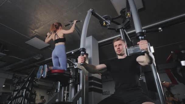남자는 가슴 근육을 위해 시뮬레이터로 운동을 한다. 한 젊은 여성 이 그 의 옆에 있는 시뮬레이터에서 당기기를 하고 있습니다. 체육관에서 훈련하고 있어요. 4K — 비디오