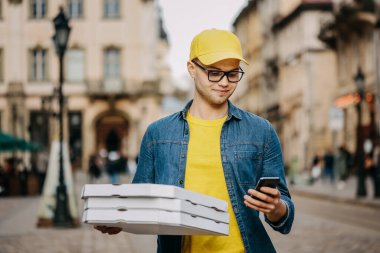 Genç bir teslimatçı üç pizza getiriyor. Gülümsüyor. Sarı bir şapka ve gözlük takıyor. Telefonuyla mesaj atıyor..