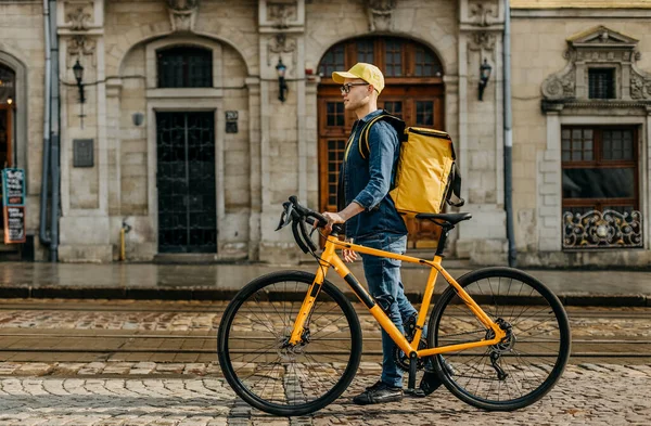 A happy delivery man is walking through the center of the old town and holding a bicycle with him. He is carrying a yellow backpack for delivery on his shoulders. Shooting from behind. — Stock Photo, Image