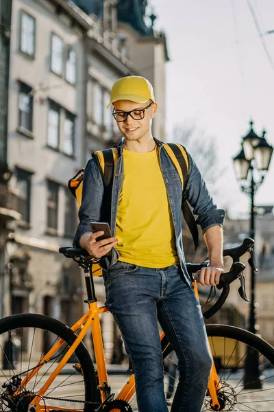 Bisikletin üzerinde oturan ve akıllı telefondan mesaj atan mutlu bir teslimatçının portresi. O, omuzlarında teslimat için sarı bir sırt çantası taşıyor. Merhaba gülümsüyor. Dikey fotoğraf.