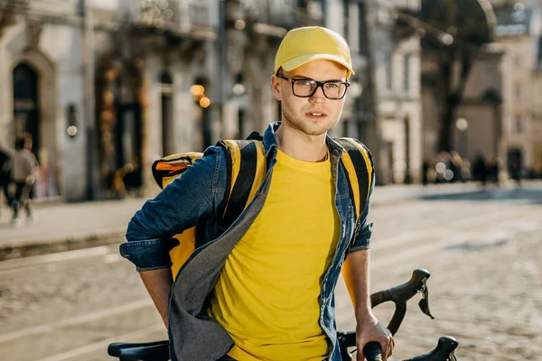 Bisiklete binip kameraya bakan mutlu bir teslimatçının portresi. O, sarı elbise giyiyor. O, omuzlarında sarı bir sırt çantası taşıyor. Eski şehrin merkezi..