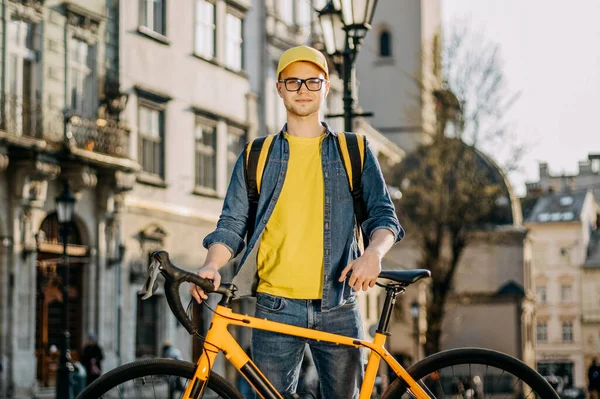 Genç bir teslimatçı ayakta ve yanında bir bisikletle duruyor. O, kameraya bakıyor. O, omuzlarında teslimat için sarı bir sırt çantası taşıyor. Eski şehrin merkezi..