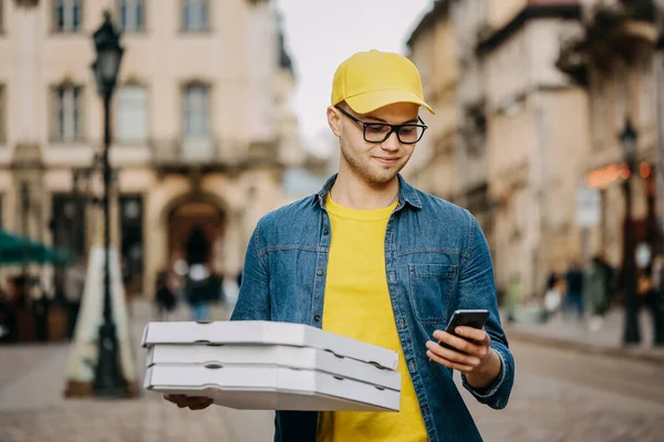 Genç bir teslimatçı üç pizza getiriyor. Gülümsüyor. Sarı bir şapka ve gözlük takıyor. Telefonuyla mesaj atıyor..