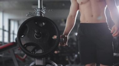 Sportif bir adam ağırlık rafına yaklaşıyor. Bir ağırlık plakasını çıkarıyor ve halterine taşıyor. Spor salonunda antrenman. 4K