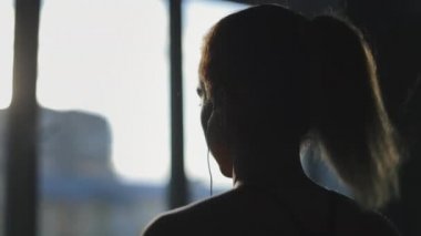 Genç bir kadın koşu bandında koşuyor ve müzik dinliyor. Pencerenin yanında koşuyor. Akşam güneşi parlıyor. Spor salonunda antrenman. Yakın plan çekim. 4K