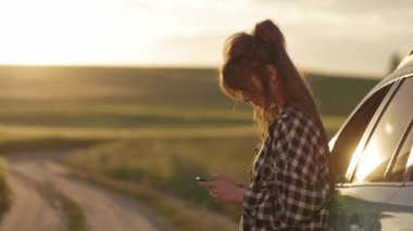 Cep telefonuyla arabanın yanında duran kadın gün batımının tadını çıkarıyor