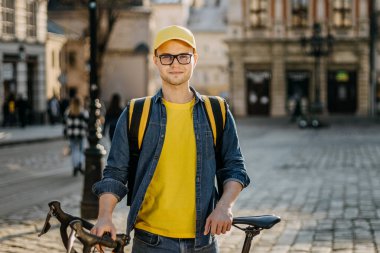 Yanında bisikleti olan genç bir teslimatçının portresi. O, kameraya bakıyor. O, omuzlarında teslimat için sarı bir sırt çantası taşıyor. Eski şehrin merkezi..