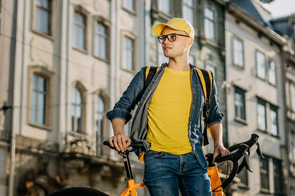 Bisiklette oturan mutlu bir teslimatçının portresi. O, sarı elbise giyiyor. O, omuzlarında sarı bir sırt çantası taşıyor. Eski şehrin merkezi..