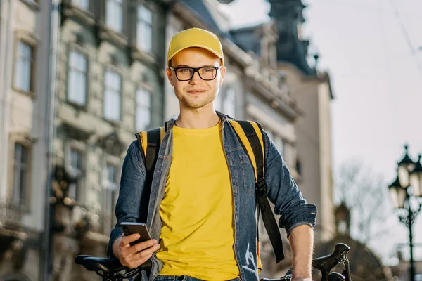Bisikletin üzerinde oturan ve akıllı telefondan mesaj atan mutlu bir teslimatçının portresi. O, omuzlarında teslimat için sarı bir sırt çantası taşıyor. Kameraya bakıyor..