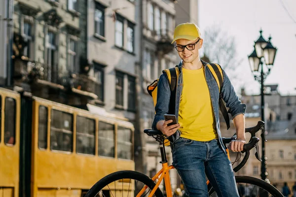Bisikletin üzerinde oturan ve akıllı telefondan mesaj atan mutlu bir teslimatçının portresi. O, kameraya bakıyor. Arka planda tramvay.