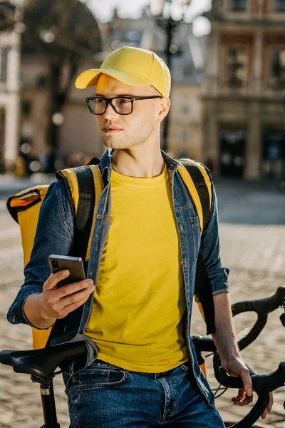 Bisikletin üzerinde oturan ve akıllı telefondan mesaj atan mutlu bir teslimatçının portresi. O, omuzlarında teslimat için sarı bir sırt çantası taşıyor. Dikey fotoğraf.