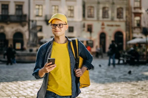 Akıllı telefondan mesaj atan mutlu bir teslimatçının portresi. Sarı bir şapka ve gözlük takıyor. Omuzlarında sarı bir sırt çantası taşıyor..