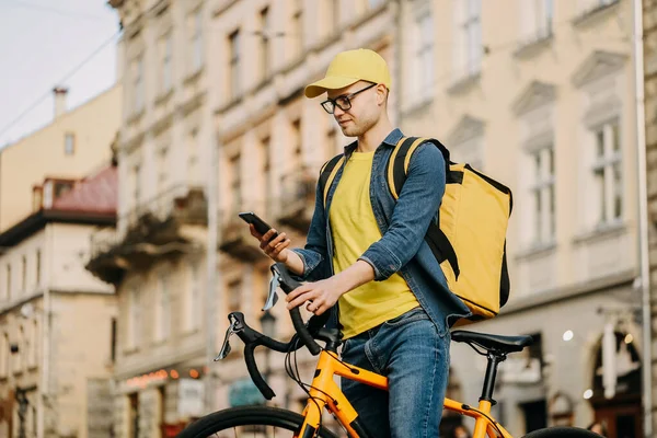 Mutlu bir teslimatçı bisiklette oturuyor ve akıllı telefonuyla mesaj atıyor. Omzunda ve şapkasında sarı bir sırt çantası taşıyor..
