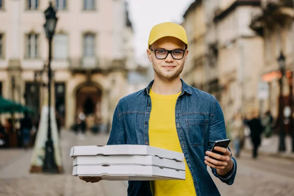 Genç, mutlu bir teslimatçı üç pizza getiriyor. Gülümsüyor. Sarı bir şapka ve gözlük takıyor. Telefonunu tutuyor ve kameraya bakıyor..