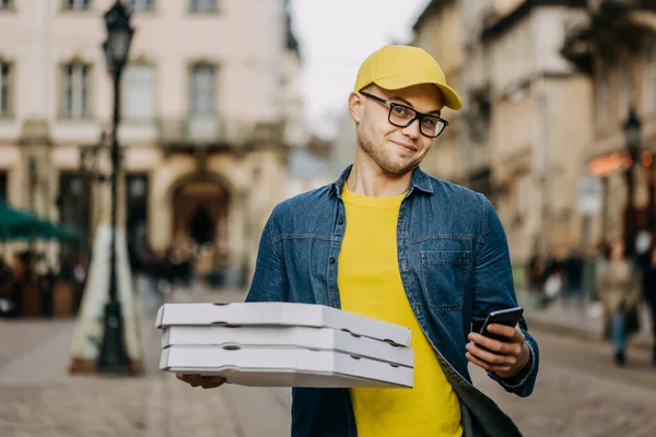 Genç, mutlu bir teslimatçı üç pizza getiriyor. Gülümsüyor. Sarı bir şapka ve gözlük takıyor. Telefonunu tutuyor ve kameraya bakıyor..