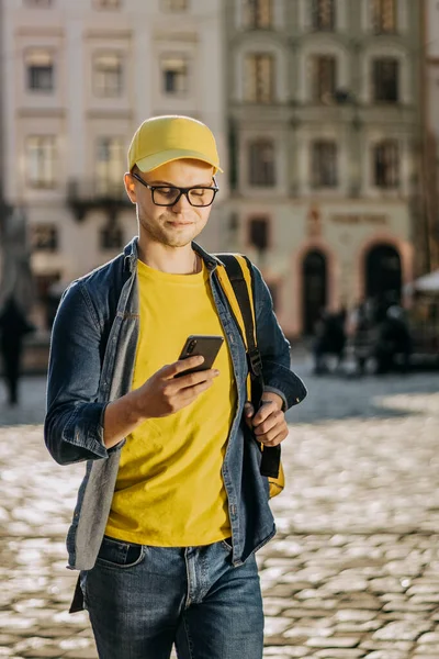 Porträtt av en glad ung förlossningsman som går och sms:ar på smartphone. Han har en gul mössa och glasögon. Han har en gul ryggsäck på axlarna. Vertikalt foto. Stockfoto